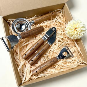 May include: A set of four kitchen utensils with wooden handles and stainless steel heads, including an egg separator, a vegetable peeler, a garlic press, and a bottle opener, all nestled in a cardboard gift box with wood shavings.