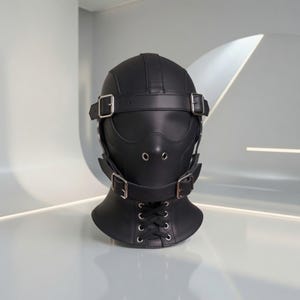 Può includere: Una maschera integrale nera con finitura liscia, dotata di fibbie in metallo argentato sulla fronte e sul mento, due aperture circolari per gli occhi, due piccoli occhielli di ventilazione nasale e un'allacciatura frontale sul collo.