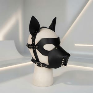 Könnte beinhalten: Eine schwarze, lederartige Tiermaske mit spitzen Ohren, einer schnauzenförmigen Nasenpartie und Augenausschnitten. Die Maske besteht aus verstellbaren Riemen, silberfarbenen Metallschnallen und Nieten und ist auf einem beigen Mannequinkopf befestigt.