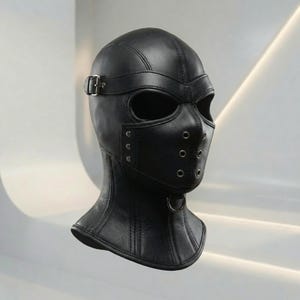 Puede incluir: Una capucha de cuero negro que cubre toda la cabeza con aberturas para los ojos, una sección para la nariz y la boca con seis ojales metálicos plateados, una correa horizontal con hebilla plateada y un anillo metálico en la barbilla.