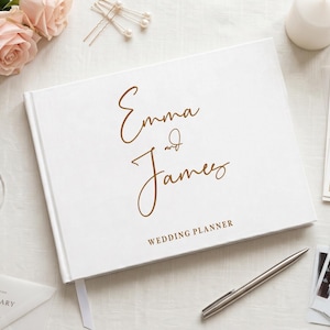 Könnte beinhalten: Ein weißes Hochzeitsplaner-Buch mit goldenem Schreibschrift-Text, der Emma and James lautet, und den darunter gedruckten Worten WEDDING PLANNER. Der Einband hat ein klares Design und das Buch liegt neben einem silbernen Stift und Schreibwaren.