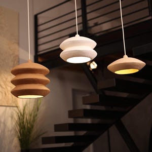 Puede incluir: Tres lámparas colgantes modernas que cuelgan del techo. Una es marrón y apilada, otra es blanca y apilada, y la tercera tiene un diseño beige en forma de platillo. Las luces cuelgan sobre una sala de estar con una escalera en el fondo.