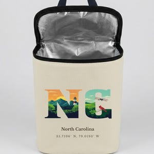 May include: Cream-colored insulated lunch bag with a black handle and zipper. The bag features the letters "NC" in a landscape design with a lighthouse, mountains, and a plane. The text "North Carolina" and coordinates are below the design.