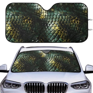 May include: A car windshield sunshade with a green and gold snakeskin pattern. The sunshade is rectangular with cutouts for the rearview mirror and is shown installed in a white car.