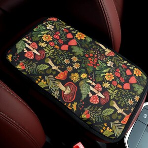 May include: A rectangular car center console armrest cover featuring a black background with a dense botanical pattern of red mushrooms, yellow flowers, green ferns, and various small leaves, finished with a black fabric border trim.