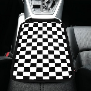 May include: Black and white checkered car console cover. The cover has a classic checkerboard pattern and a black border. The cover is designed to fit over the center console of a vehicle.