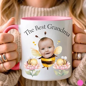 May include: White ceramic mug with a pink interior and handle. The mug features the text "The Best Grandma" above a baby dressed as a bee, with two cartoon bees and flowers. The mug is held by a person.