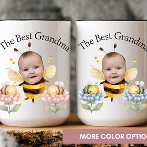 May include: Two white ceramic mugs with black handles and rims. Each mug features a baby's face with bee wings and a bee costume, surrounded by flowers and the text "The Best Grandma".