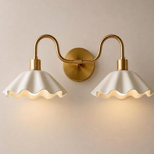 May include: A double wall sconce featuring a circular brass backplate and two curved brass arms extending to support two white, scalloped, bell-shaped shades. The fixture emits a soft, warm glow from beneath each of the two fluted ceramic shades.