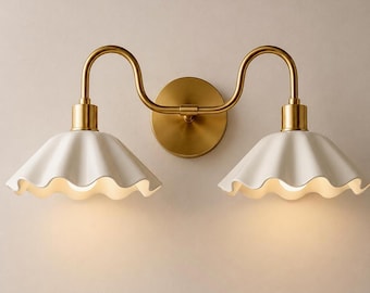 Può includere: Una lampada da parete doppia con una piastra circolare in ottone e due bracci curvi in ottone che sostengono due paralumi bianchi a forma di campana con bordi smerlati. L'apparecchio emette una luce calda e soffusa sotto ogni paralume scanalato.