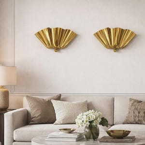 May include: Two gold-colored wall sconces with a fan-like design are mounted on a white wall. The sconces are above a cream-colored sofa with decorative pillows. A lamp and side table are on the left.