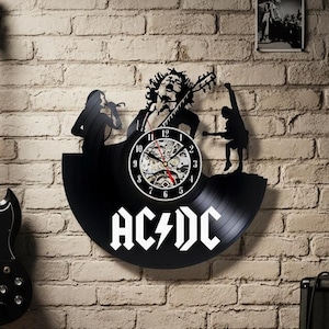 Könnte beinhalten: Eine schwarze E-Gitarre hängt an einer Backsteinmauer neben einer schwarzen Vinyl-Schallplattenuhr mit dem Bandnamen "AC/DC" in Weiß. Ein schwarzer Marshall-Verstärker steht auf dem Boden. Schallplatten und ein gerahmtes Foto sind ebenfalls zu sehen.