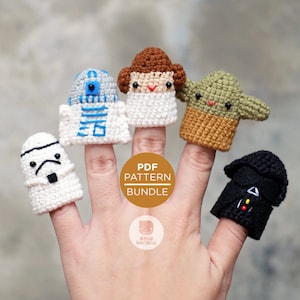 May include: A set of five crochet finger puppets featuring characters from Star Wars. The puppets are R2-D2, Princess Leia, Yoda, Darth Vader, and Stormtrooper. The puppets are all white, except for Yoda, who is green, and Darth Vader, who is black. The puppets are all wearing their iconic costumes. The image also includes a gold circle with the text "PDF PATTERN BUNDLE".