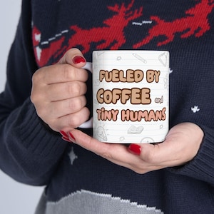 May include: White ceramic coffee mug with the text "Fueled by Coffee and Tiny Humans" in brown and black. The mug has a lined paper design with small illustrations of a pencil, lightbulb, and book.