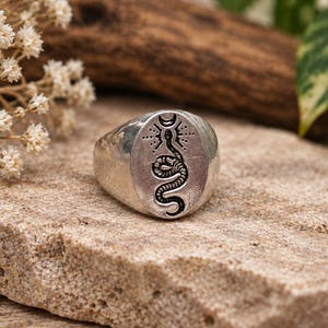 May include: A silver-toned metal ring featuring an oval face engraved with a black-filled snake design. Above the snake is a crescent moon symbol with radiating lines. The ring has a smooth, polished band and a weathered, textured finish.