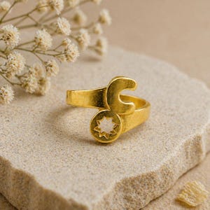 Pode incluir: Um anel dourado com uma chave inglesa e um design circular com um recorte em forma de estrela. O anel é colocado sobre um fundo neutro com pequenas flores brancas, criando uma estética simples e elegante. Uma peça de joalharia única.