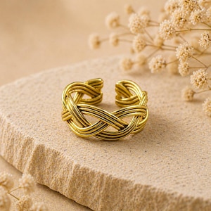 May include: A gold-colored adjustable ring with a woven design. The ring is open at the top and features a series of interwoven lines, creating a textured appearance. The ring is displayed on a light brown stone surface, with dried flowers in the background.