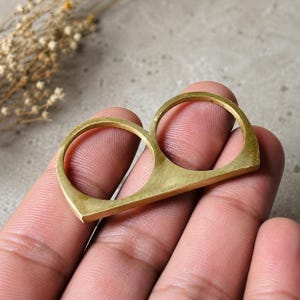 May include: A two-finger ring crafted from brushed brass, featuring two circular openings for fingers connected by a flat, horizontal bar. The metal has a matte, golden-toned finish with visible fine grain texture across the surface.