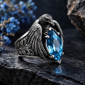 May include: A silver ring featuring a detailed raven with spread wings clutching a large, marquise-cut, bright blue gemstone. The band is decorated with intricate, embossed leaf patterns, and the metal has an oxidized finish to highlight the texture.