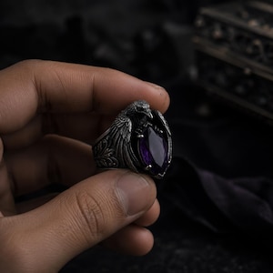 May include: A silver ring with a raven motif, its wings framing a large, oval, deep purple gemstone. The ring showcases detailed floral and feather patterns. The raven's head is positioned above the gemstone.
