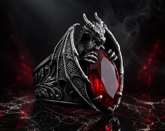May include: A silver-toned ring featuring a sculpted dragon with spread wings clutching a large, faceted, marquise-cut red gemstone. The band displays intricate, embossed leaf-like patterns along the sides, finished with a dark, oxidised metallic texture.