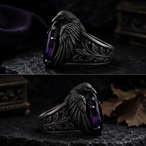 May include: A silver ring with a raven design, holding a large, oval-cut purple gemstone. The ring has detailed carvings on the band and a dark, antique finish. The raven's wings are spread, and the gemstone is a deep violet colour. The ring is a statement piece.