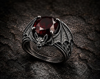 May include: A silver-toned ring with a bat motif and a large, oval-cut, deep red gemstone. The ring's design incorporates bat wings and a bat's face. The ring has a gothic style, with intricate detailing, and is suitable for a variety of occasions.