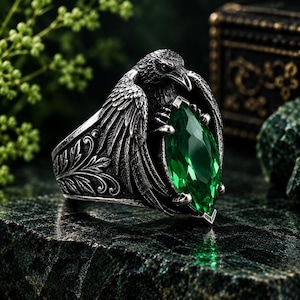 May include: A silver-toned ring featuring a detailed raven with spread wings clutching a large, marquise-cut emerald green gemstone. The band is decorated with intricate leaf-like engravings, and the metal has a dark, oxidized finish.
