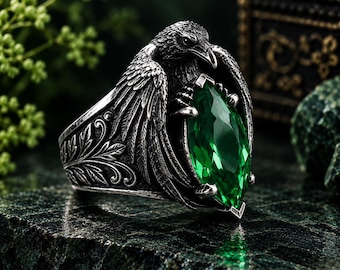 May include: A silver-toned ring featuring a detailed raven with spread wings clutching a large, marquise-cut emerald green gemstone. The band is decorated with intricate leaf-like engravings, and the metal has a dark, oxidized finish.