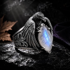 May include: A silver ring featuring a detailed raven with spread wings clutching a marquise-cut moonstone. The band is decorated with intricate leaf-like engravings, and the metal has a dark, oxidized finish that highlights the bird's feathers.