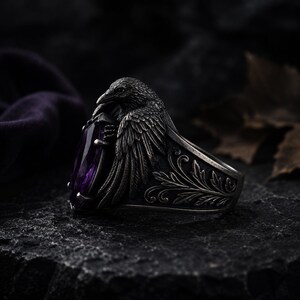 May include: A silver ring with a raven motif, holding a large, purple, teardrop-shaped gemstone. The ring has detailed feather and floral patterns. The background is dark and textured.