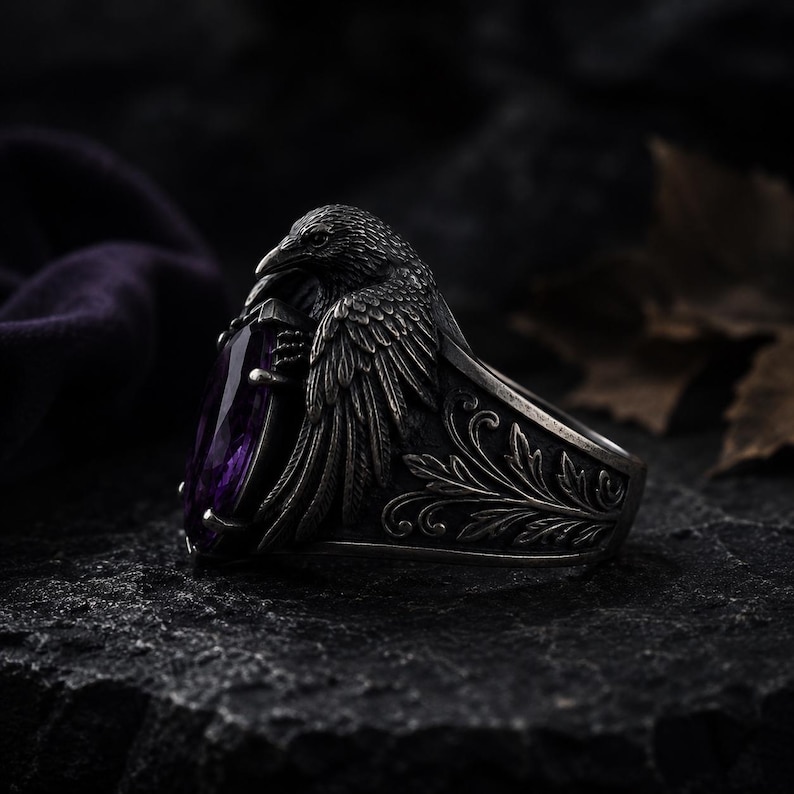 May include: A silver ring with a raven motif, holding a large, purple, teardrop-shaped gemstone. The ring has detailed feather and floral patterns. The background is dark and textured.