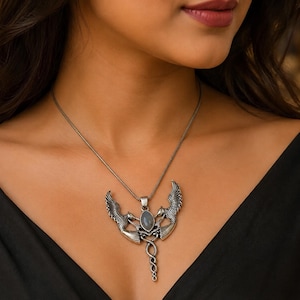 Puede incluir: Un collar con colgante de metal plateado que presenta dos aves simétricas con las alas extendidas flanqueando una piedra gris ovalada central. Las aves están situadas sobre un tallo de metal retorcido con forma de serpiente.
