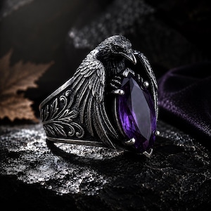 May include: A silver ring with a raven design, holding a purple gemstone. The ring has an ornate band with a floral pattern. The raven's wings are spread, and the gemstone is a large, marquise-cut. The ring has a gothic or fantasy theme.