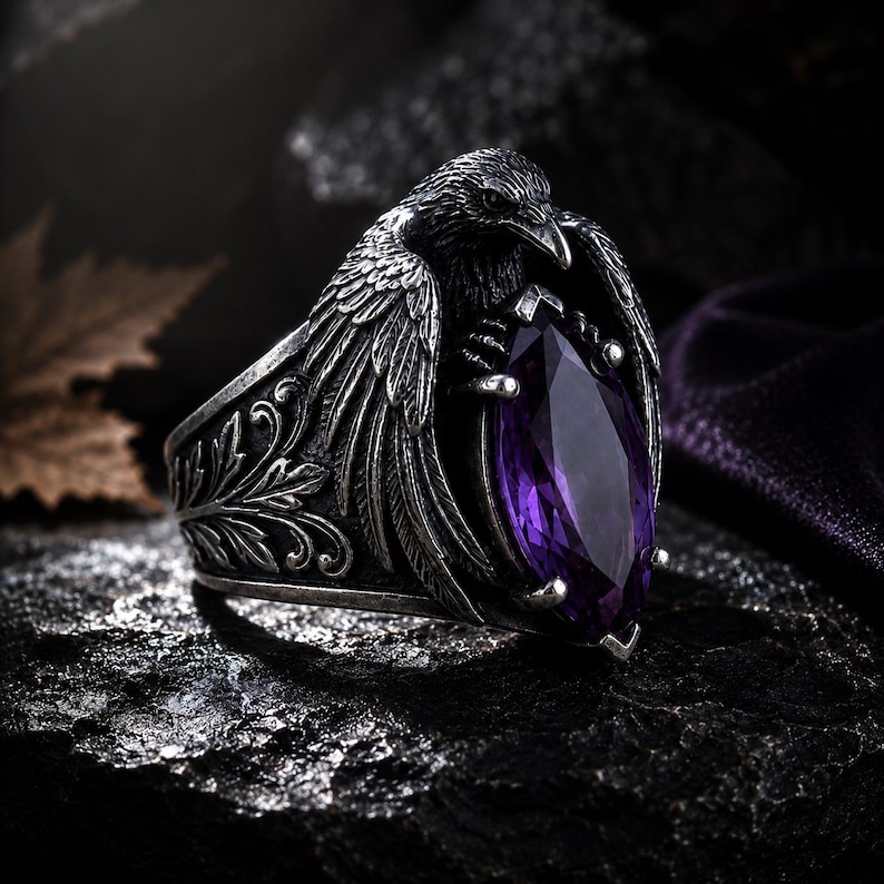 May include: A silver ring with a raven design, holding a purple gemstone. The ring has an ornate band with a floral pattern. The raven's wings are spread, and the gemstone is a large, marquise-cut. The ring has a gothic or fantasy theme.