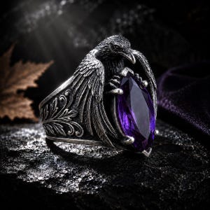 May include: A silver-toned ring featuring a detailed raven design holding a large, marquise-cut purple gemstone. The ring has intricate feather and floral detailing, set against a dark, textured background.