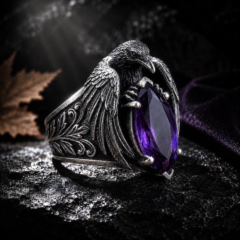 May include: A silver ring with a raven motif, holding a large, purple, marquise-cut gemstone. The ring has detailed feather and floral patterns. The background is dark and textured.
