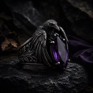May include: A silver ring with a raven motif, its wings embracing a large, purple, teardrop-cut gemstone. The ring has an engraved band with an antique finish. The raven's detailed feathers and the gemstone's deep colour are set against a dark backdrop.