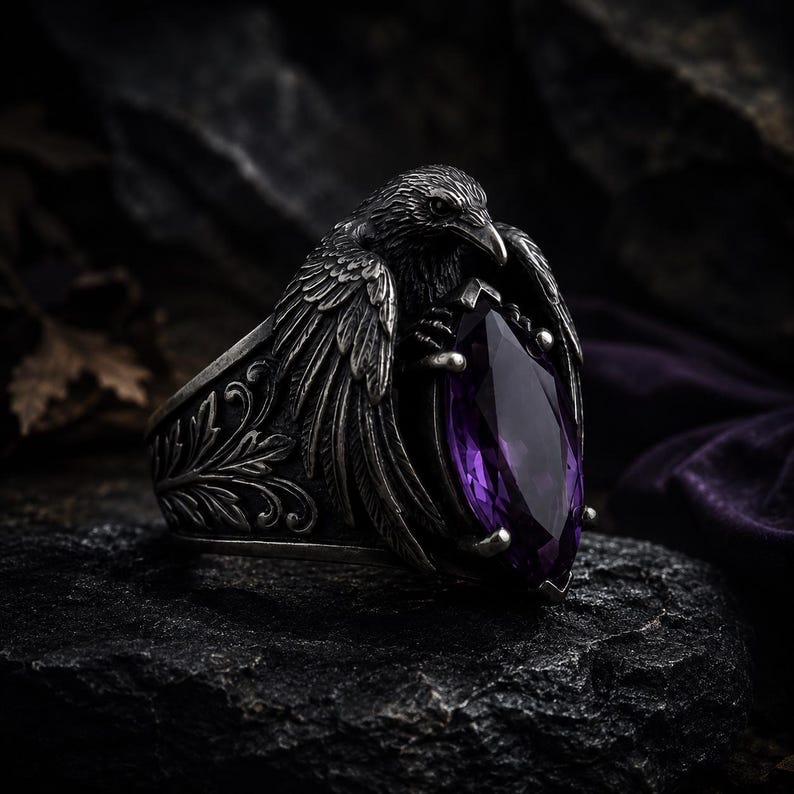 May include: A silver ring with a raven motif, its wings embracing a large, purple, teardrop-cut gemstone. The ring has an engraved band with an antique finish. The raven's detailed feathers and the gemstone's deep colour are set against a dark backdrop.