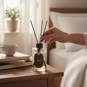 May include: A clear glass diffuser bottle with black reeds, labeled "Yin". The bottle sits on a wooden nightstand next to a stack of books and a teacup. A hand is adjusting the reeds. The scene is set in a bedroom.