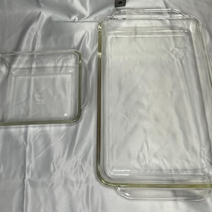 May include: Two clear glass baking dishes. One is rectangular with handles, the other is square with handles. Both are empty and sit on a white surface. The dishes are made of clear glass and are designed for baking.