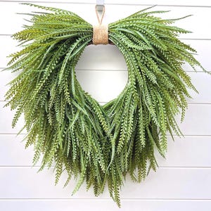 May include: A round, green artificial fern wreath. The wreath is made of many small, delicate leaves and is tied together with a natural twine. It hangs against a white, horizontal-slat background.