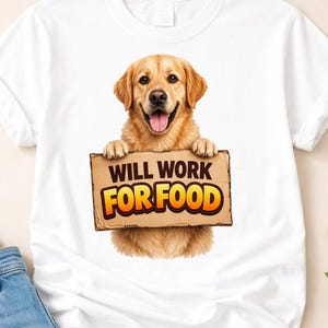 Può includere: T-shirt bianca con un golden retriever che tiene in mano un cartello con la scritta "WILL WORK FOR FOOD" in lettere arancioni e gialle. Il cane ha un'espressione felice. La maglietta è abbinata a jeans blu.