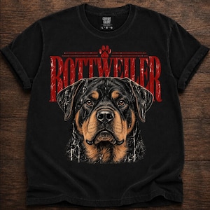 May include: A black short-sleeved t-shirt featuring a graphic of a Rottweiler dog's face in the center. Above the dog, the word ROTTWEILER is printed in large, distressed red capital letters, with a small red paw print icon centered above the text.