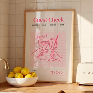 May include: A framed pink and white "Guest Check" print with the word "cheers!" and illustrations of two hands toasting glasses. The print has a wooden frame and is displayed in a kitchen setting. A bowl of lemons sits in front of the print.