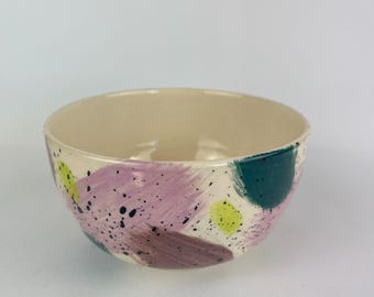 Może przedstawiać: Ceramiczna miska z kremowym wnętrzem i zewnętrzem, ozdobiona abstrakcyjnymi pociągnięciami pędzla. Na zewnątrz znajdują się plamy lawendy, turkusu, brązu i żółtej farby, a także czarne plamki. Miska ma zaokrąglony kształt.
