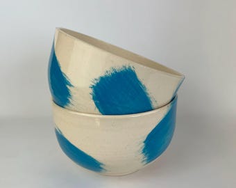 Może przedstawiać: Dwie ceramiczne miski ułożone jedna na drugiej. Miski są w kolorze złamanej bieli z odważnymi, niebieskimi pociągnięciami pędzla. Miski mają prosty, nowoczesny design, odpowiedni do serwowania jedzenia lub jako elementy dekoracyjne.