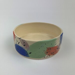 May include: A ceramic bowl with a cream interior and a colourful exterior. The exterior is decorated with blue, green, orange, and purple brushstrokes, along with black speckles. The bowl is round and has a low profile.