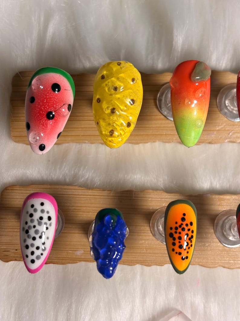 May include: A selection of fruit-themed nail art designs. The nails are decorated with watermelon, pineapple, and other fruit designs. Each nail is painted with vibrant colours and intricate details, showcasing a playful and artistic style.