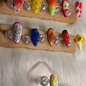 May include: Artificial nails with fruit-themed designs, such as watermelon, pineapple, and dragon fruit. The nails are painted in bright colours and displayed on wooden holders. The nail art features detailed designs and glossy finishes.